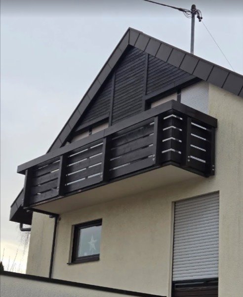 Balkon mit dunkel lasierter Holzverkleidung und waagerechten Geländerbrettern an einer Dachgaube.