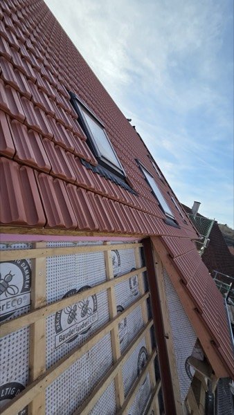 Rotes Ziegeldach mit mehreren Dachflächenfenstern, darunter eine Giebelwand mit Holzlattung während der Bauarbeiten.