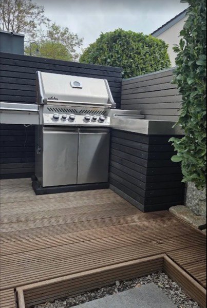 Outdoorküche mit Gasgrill vor einer schwarz gestrichenen Sichtschutzwand aus Holz, davor eine Holzterrasse.